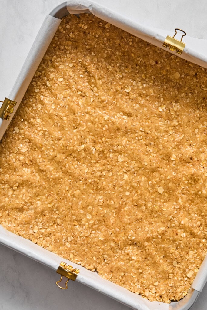 A square baking pan lined with parchment paper, filled with an unbaked oat and dough mixture featuring creamy almond butter, ready to be baked into delicious vegan bars.