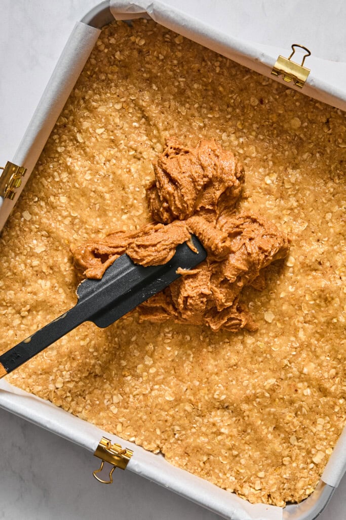 A baking pan lined with parchment paper holds an oat mixture. A spatula is spreading a layer of creamy almond butter filling on top, creating irresistible vegan jelly bars.