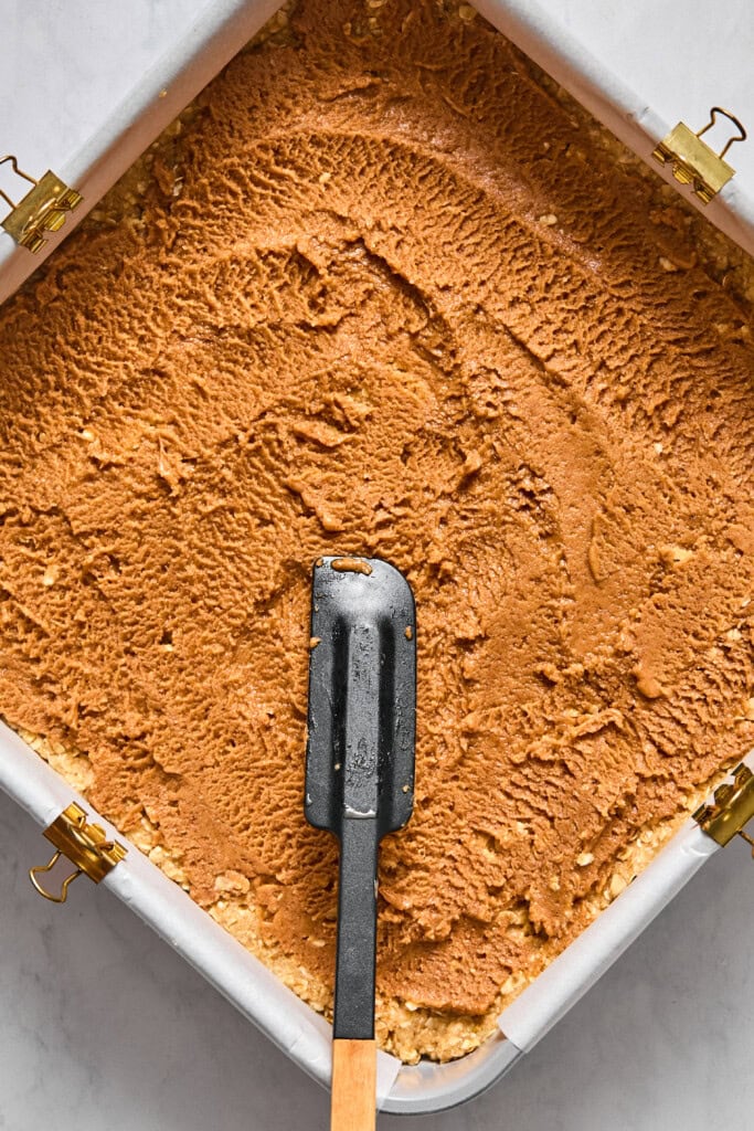 A square pan filled with a smooth brown almond butter mixture, partially spread with a black spatula, lined with parchment paper and clipped at the sides. Ideal for making delicious vegan jelly bars.