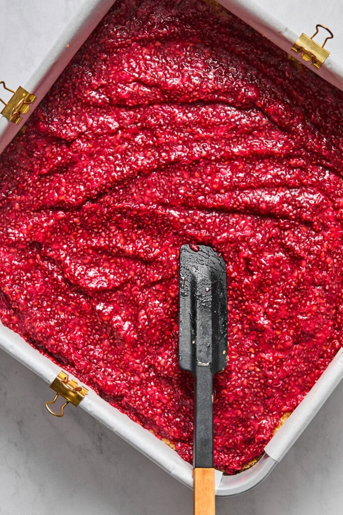 A square baking pan filled with a bright red vegan raspberry mixture, smoothed with a black spatula with a wooden handle, sits on a light surface-perfect for making delicious jelly bars.