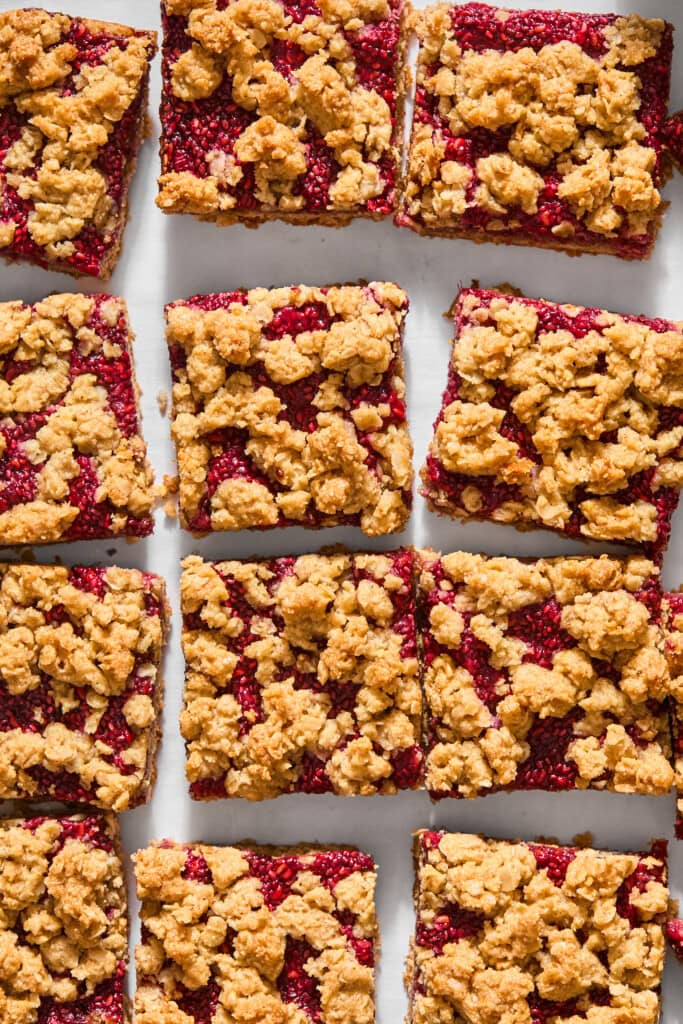Twelve vegan almond butter and jelly bars with a golden oat topping are arranged in a grid pattern on a white surface.