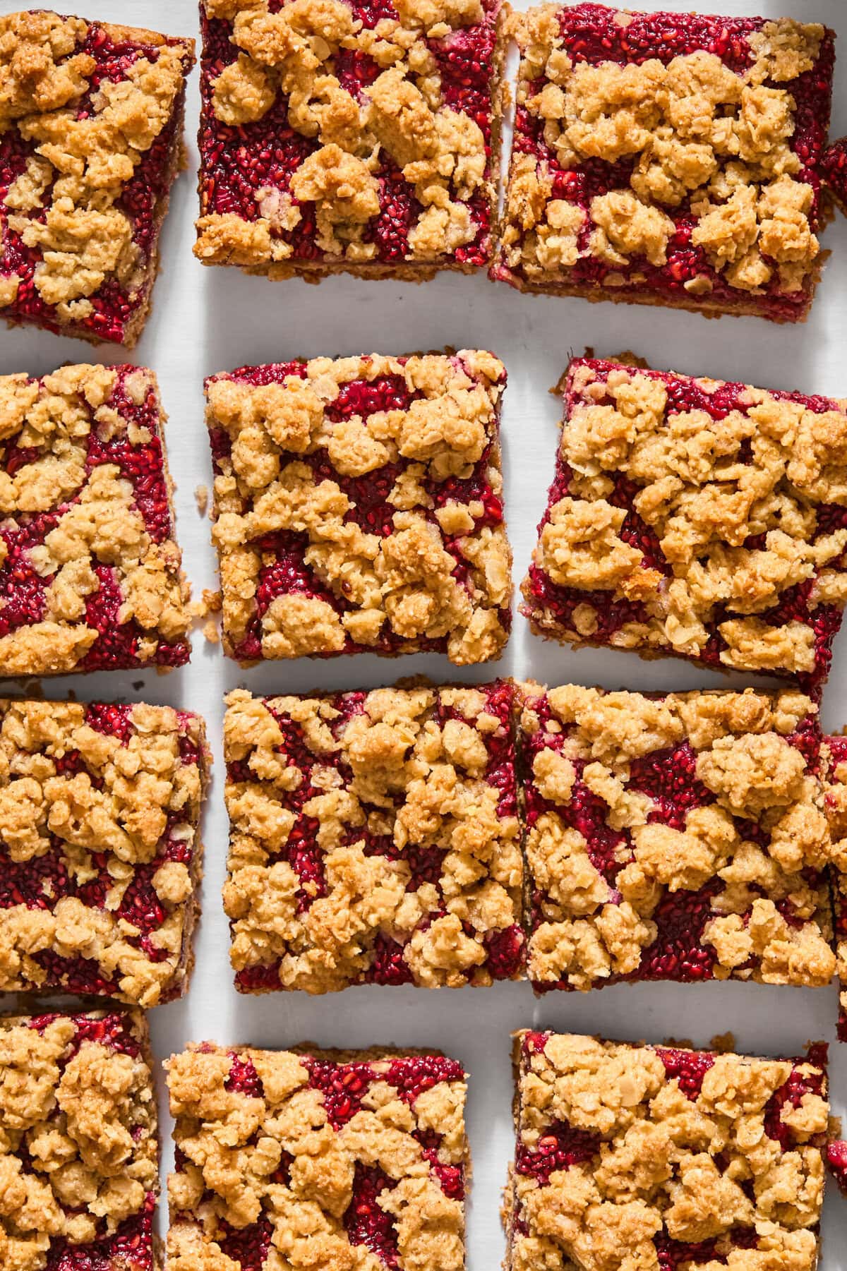 Twelve vegan almond butter and jelly bars with a golden oat topping are arranged in a grid pattern on a white surface.