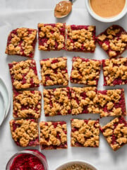 Sixteen almond butter and jelly bars are arranged on parchment paper, surrounded by bowls of oats, raspberry jam, peanut butter, and plates. A spoon with peanut butter rests beside the bars.