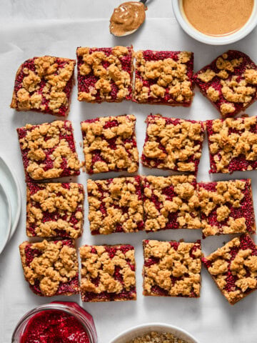 Sixteen almond butter and jelly bars are arranged on parchment paper, surrounded by bowls of oats, raspberry jam, peanut butter, and plates. A spoon with peanut butter rests beside the bars.