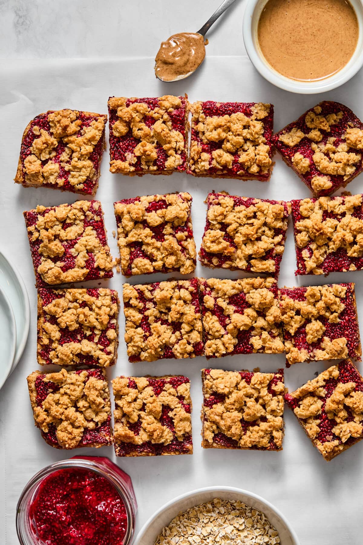 Sixteen almond butter and jelly bars are arranged on parchment paper, surrounded by bowls of oats, raspberry jam, peanut butter, and plates. A spoon with peanut butter rests beside the bars.