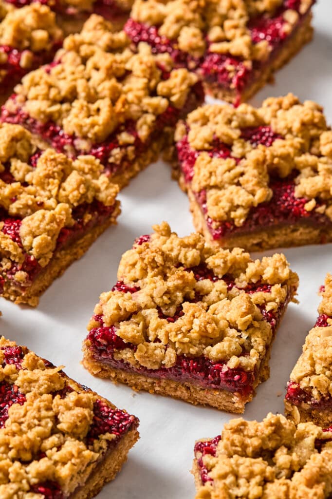 Close-up of several vegan oat crumble bars with a raspberry filling, arranged on a white parchment surface. The bars have a golden-brown crumbly topping and visible red fruit layer, reminiscent of classic jelly bars.