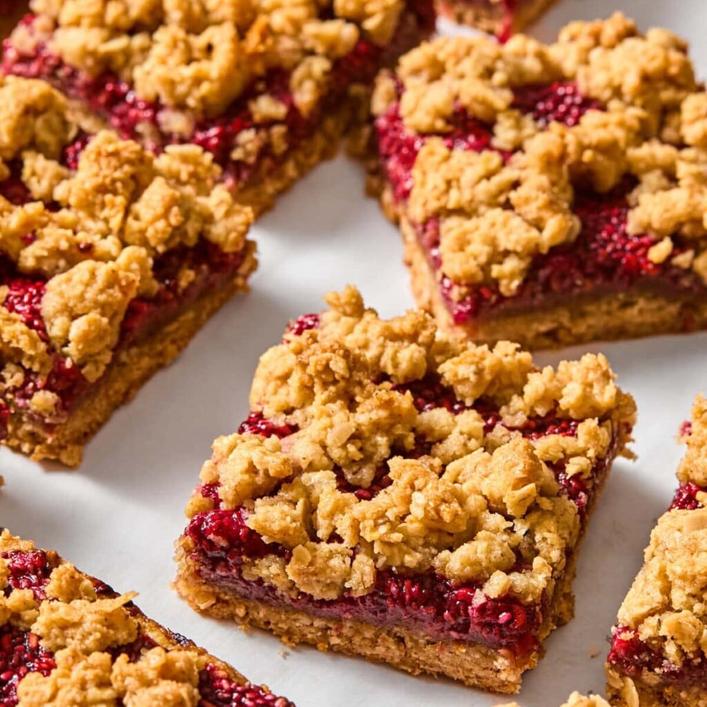 Close-up of several vegan almond butter and jelly bars with a raspberry filling, arranged on a white parchment surface. The bars have a golden-brown crumbly topping and visible red fruit layer, reminiscent of classic jelly bars.