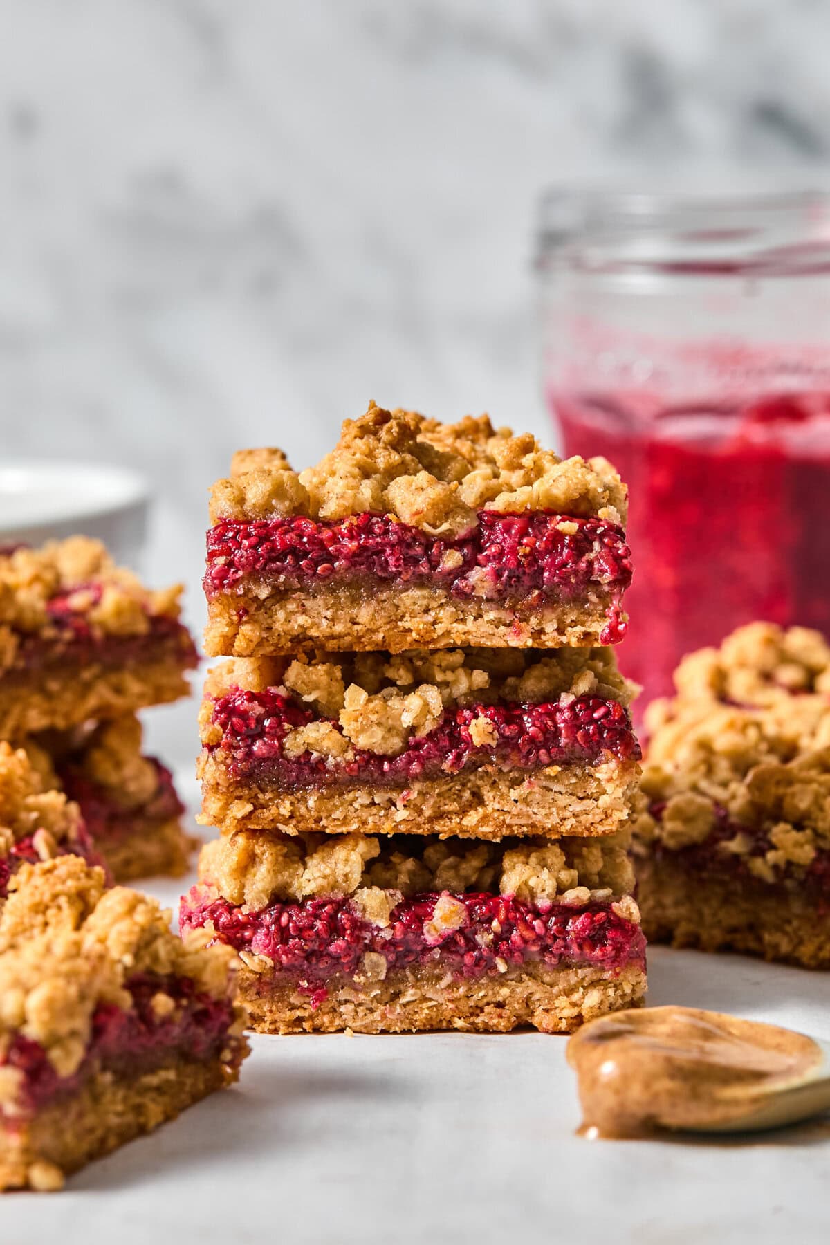 Three stacked almond butter & jelly bars with crumbly layers, set on a white surface. Made with wholesome ingredients like almond butter, these vegan and gluten-free bars are perfect for a guilt-free treat. A glass jar filled with raspberry jam is blurred in the background. Other bars and a spoon are also visible.