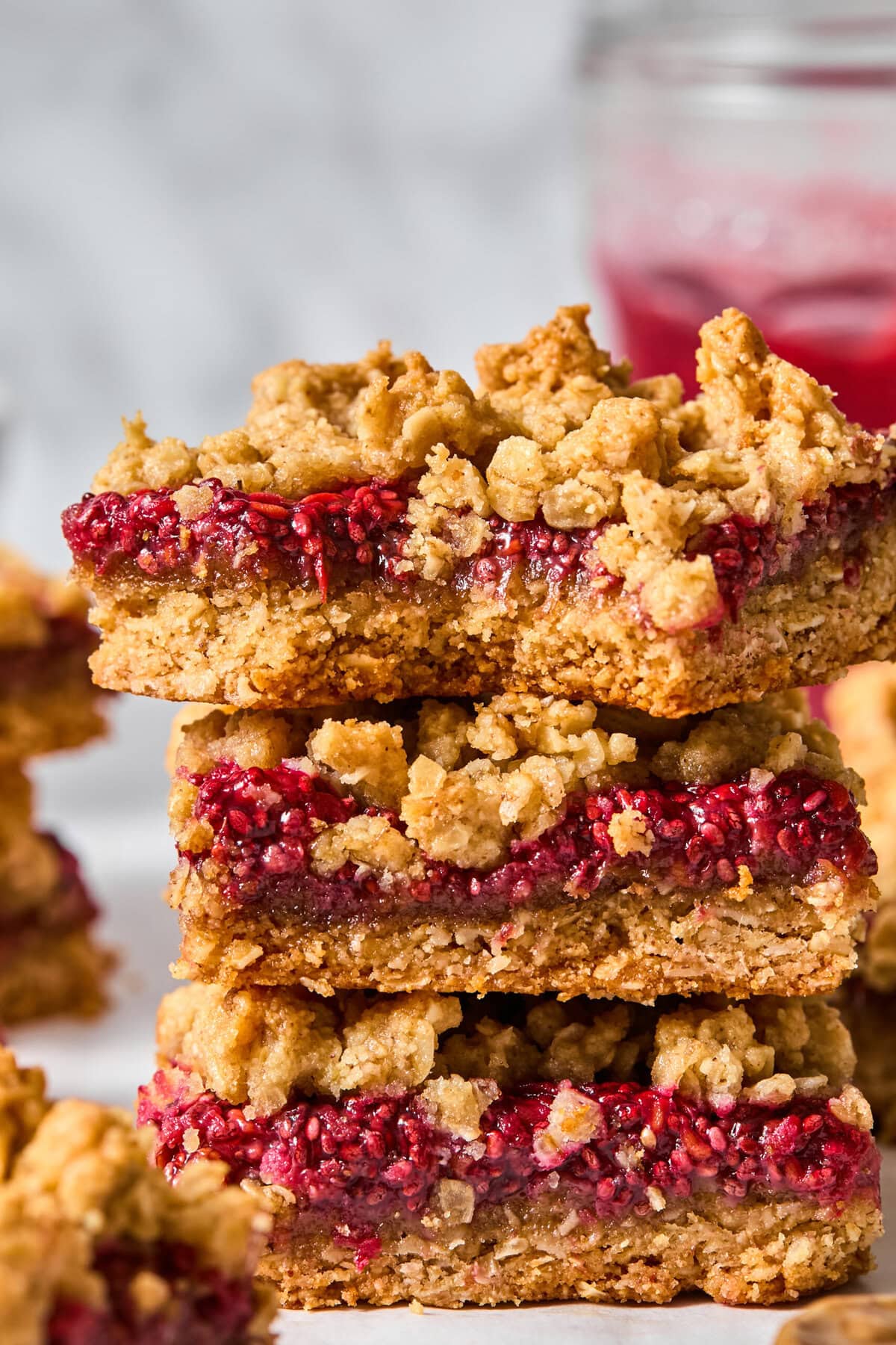 Three raspberry oat crumble bars stacked on top of each other, showing layers of golden oat crust and bright red raspberry filling—reminiscent of classic almond butter and jelly bars. A glass jar with a red drink is blurred in the background.
