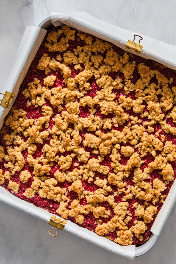 A square baking pan filled with vegan raspberry crumble bars, featuring a golden crumb topping over a vibrant red fruit layer, lined with parchment paper.