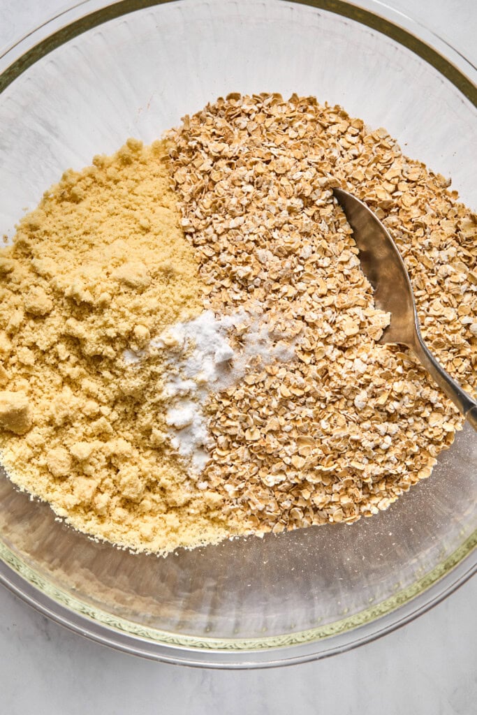 A glass bowl containing almond flour, rolled oats, baking powder, and a spoon on a light surface-perfect ingredients for whipping up vegan treats like almond butter jelly bars.