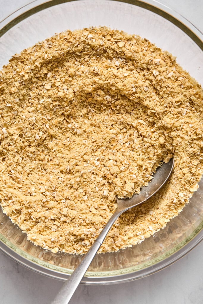 A metal spoon sits in a glass bowl filled with a dry, crumbly mixture of oats and flour-likely the vegan base for almond butter jelly bars-seen from directly above.