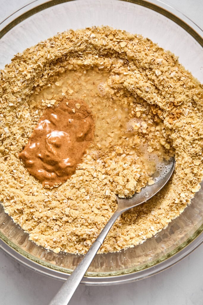 A glass bowl with oats, liquid, and a dollop of almond butter being mixed with a metal spoon, ready to be combined for baking vegan treats or jelly bars.