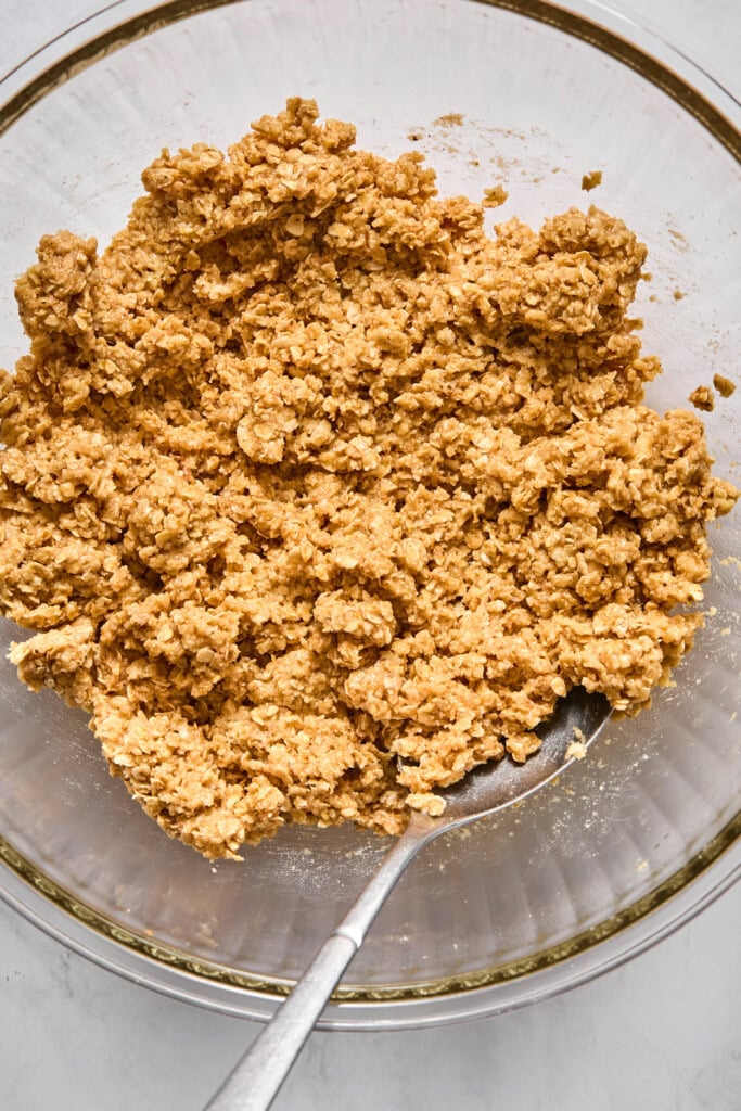 A glass bowl filled with a mixture of oats and a light brown almond butter batter, with a metal spoon resting inside, ready for vegan cookie or jelly bar preparation.