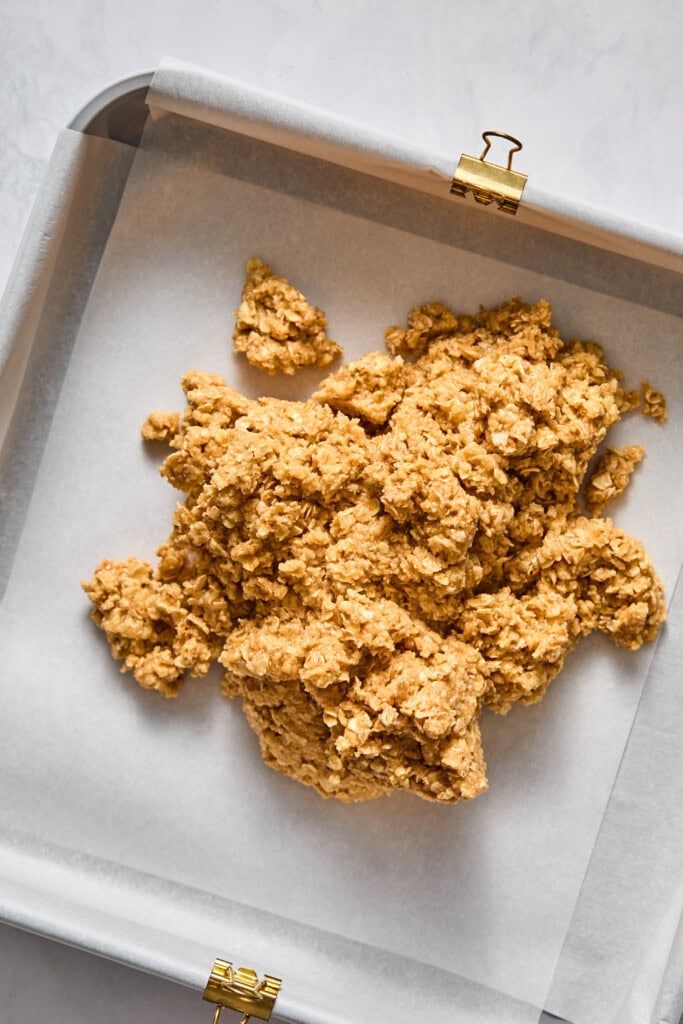 A baking tray lined with parchment paper holds a pile of unbaked, chunky oat cookie dough infused with rich almond butter, ready to be spread or shaped into irresistible vegan treats or jelly bars before baking.