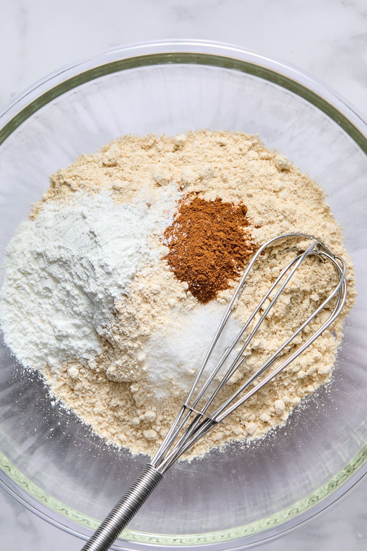 A glass bowl contains flour, baking powder, cinnamon, and salt, with a metal whisk resting inside, ready for mixing fluffy pancakes.