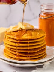 A stack of fluffy pumpkin pancakes topped with butter is drizzled with syrup. A jar of orange spread and a fork are nearby, on a white plate and napkin, with a marble background.