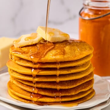 A stack of fluffy pumpkin pancakes topped with butter is drizzled with syrup. A jar of orange spread and a fork are nearby, on a white plate and napkin, with a marble background.