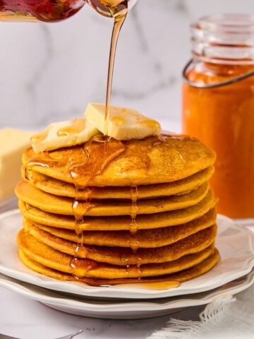 A stack of fluffy pumpkin pancakes topped with butter is drizzled with syrup. A jar of orange spread and a fork are nearby, on a white plate and napkin, with a marble background.