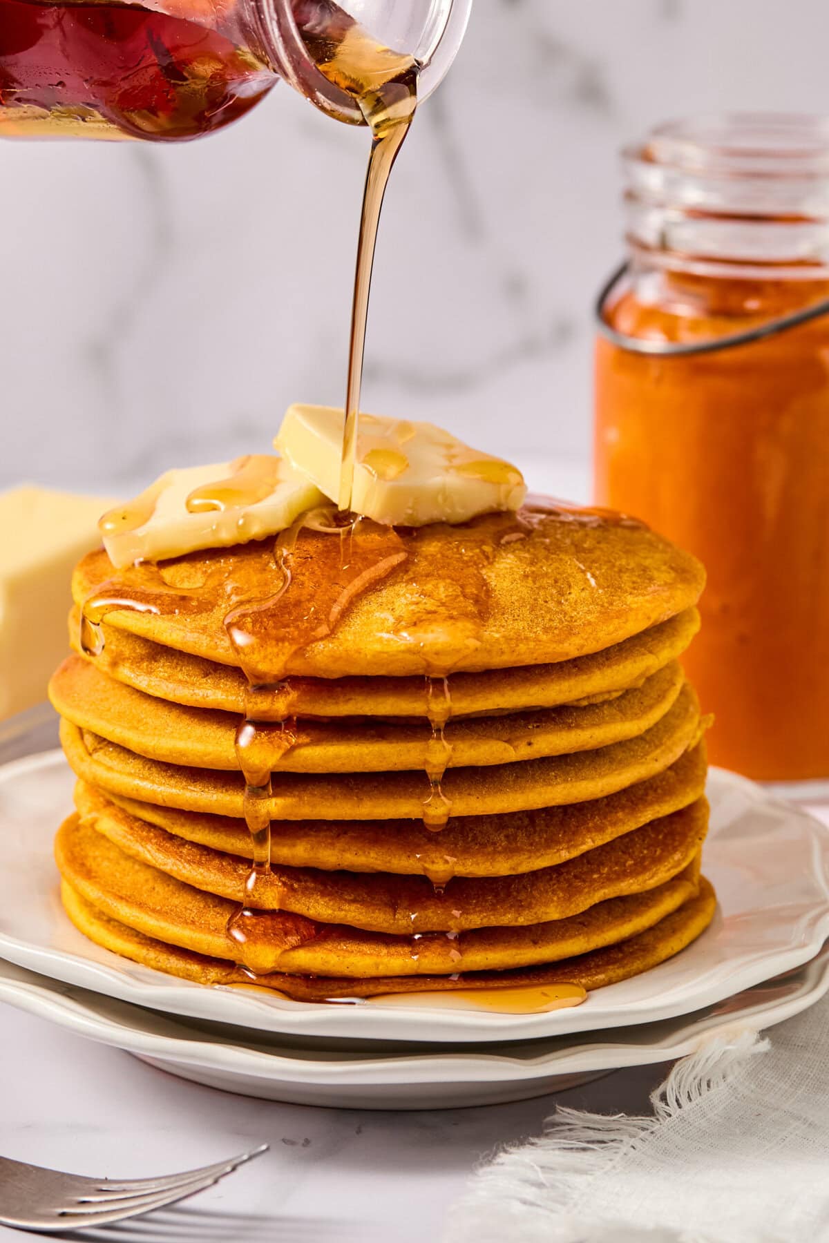 A stack of fluffy pumpkin pancakes topped with butter is drizzled with syrup. A jar of orange spread and a fork are nearby, on a white plate and napkin, with a marble background.