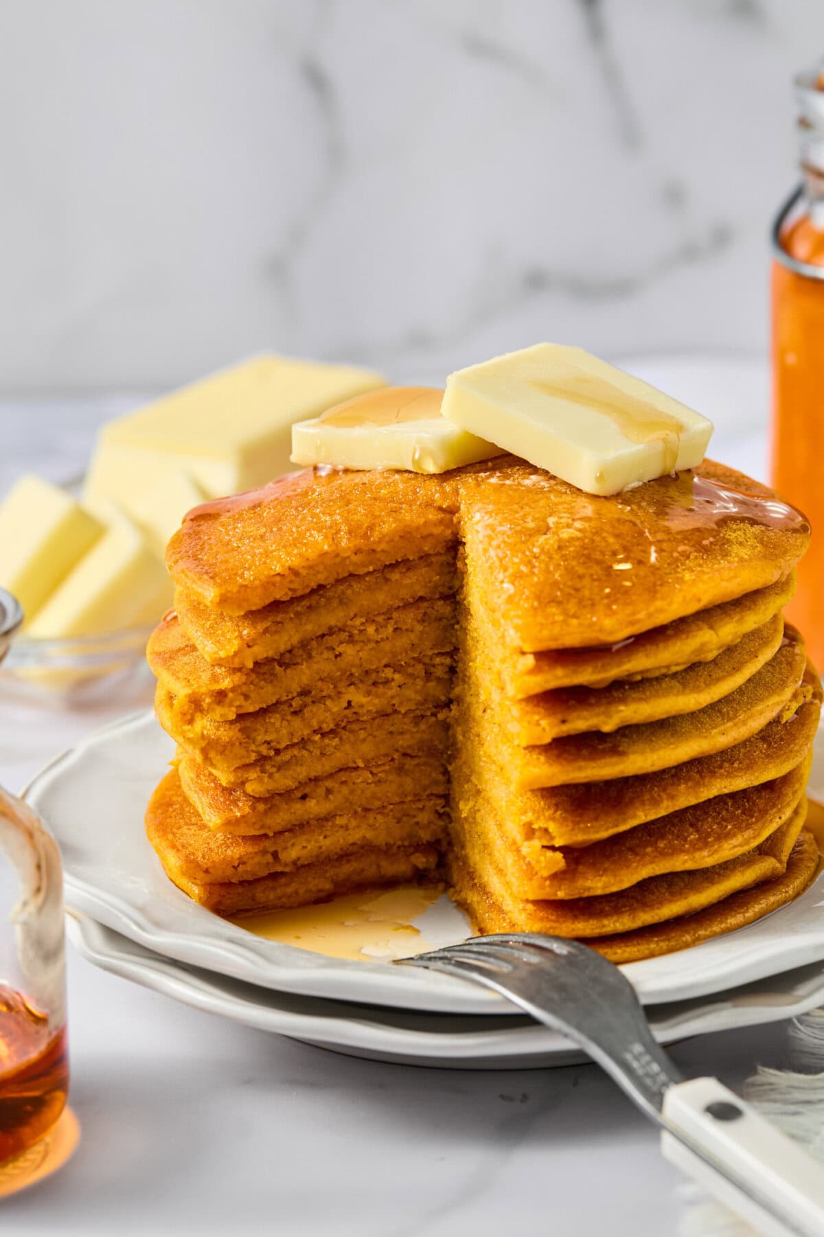 A stack of fluffy pancakes with a slice cut out, topped with butter and syrup, sits on a plate. Perfect for cozy mornings, these Vegan Pumpkin Pancakes are surrounded by a fork in front and blocks of butter and a honey jar in the background on a white surface.
