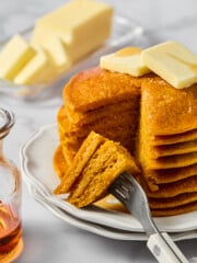 A stack of fluffy pancakes with butter on top sits on a white plate. A fork holds a cut bite. A glass syrup jar and a dish of butter are in the background, all on a marble surface.