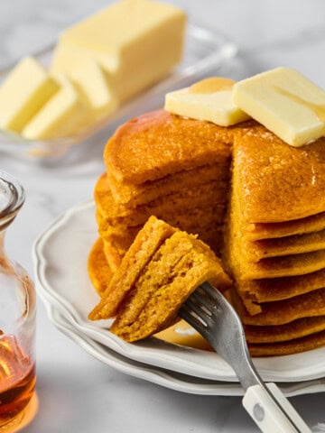 A stack of fluffy pancakes with butter on top sits on a white plate. A fork holds a cut bite. A glass syrup jar and a dish of butter are in the background, all on a marble surface.