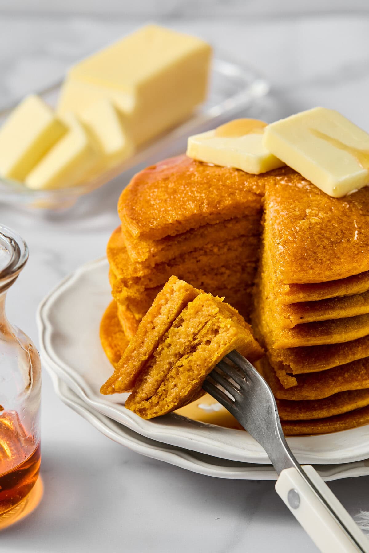 A stack of fluffy pancakes with butter on top sits on a white plate. A fork holds a cut bite. A glass syrup jar and a dish of butter are in the background, all on a marble surface.