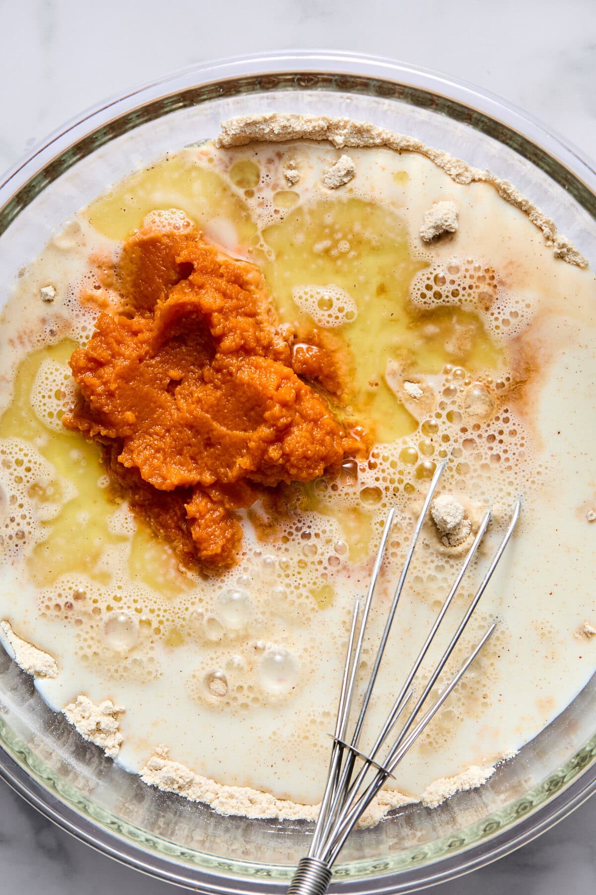 A glass bowl with flour, milk, eggs, oil, and pumpkin puree being mixed with a metal whisk on a marble countertop-ideal for creating fluffy pancakes or delicious vegan pumpkin pancakes.