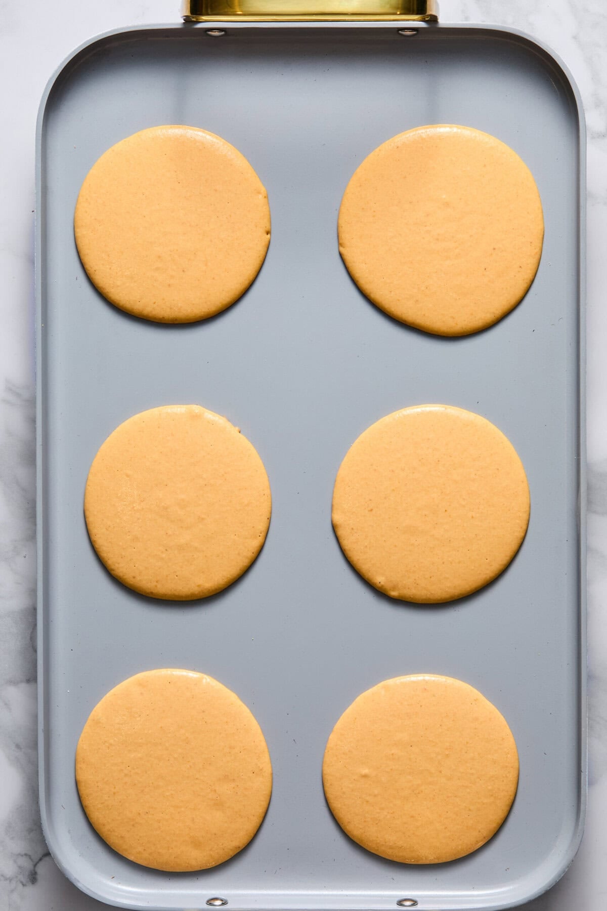Six round, evenly spaced fluffy pancake batters sit on a rectangular non-stick griddle, ready to be cooked. The griddle rests atop a marble countertop, perfectly prepped for making delicious Vegan Pumpkin Pancakes or Gluten-Free breakfast treats.