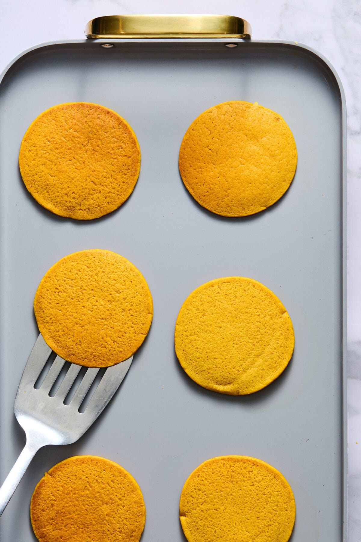 A gray baking sheet with six golden brown, fluffy pancakes; a metal spatula is lifting one Vegan Pumpkin Pancake from the lower left.