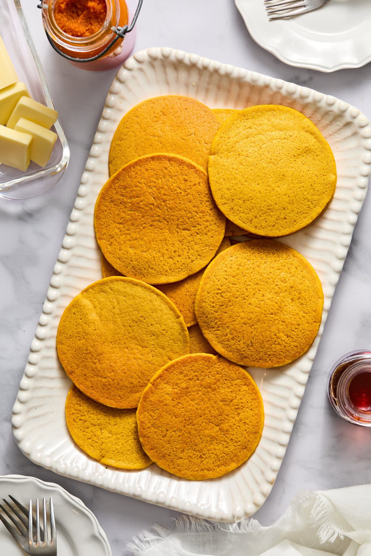 A white platter holds eight golden, fluffy pancakes. Surrounding the platter are sticks of butter, a jar of jam, a glass of red juice, plates, and forks, all set on a white marble surface-an inviting setup perfect for serving delicious Vegan Pumpkin Pancakes.