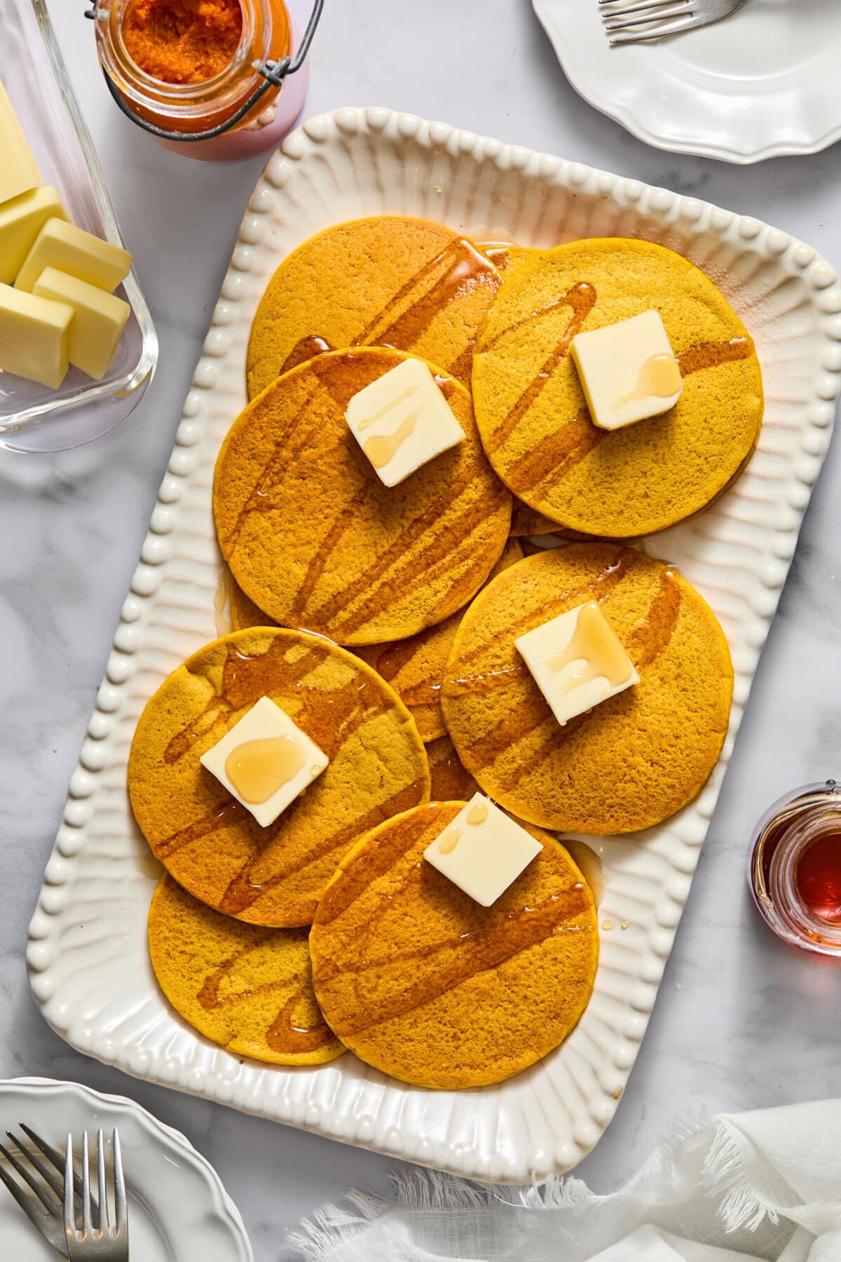 A rectangular white platter with seven fluffy pancakes topped with butter pats and drizzled syrup, surrounded by jars of syrup, butter, forks, and plates on a marble surface—perfect for serving gluten-free pancakes or vegan pumpkin pancakes at your next brunch gathering.