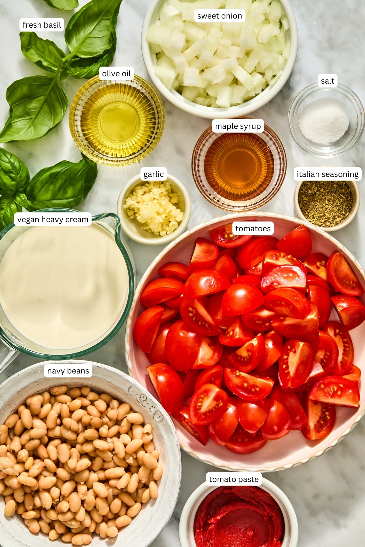 Ingredients for creamy Tuscan white beans arranged on a counter: fresh basil, diced onion, olive oil, salt, maple syrup, minced garlic, Italian seasoning, vegan heavy cream, halved cherry tomatoes, navy beans, and tomato paste.