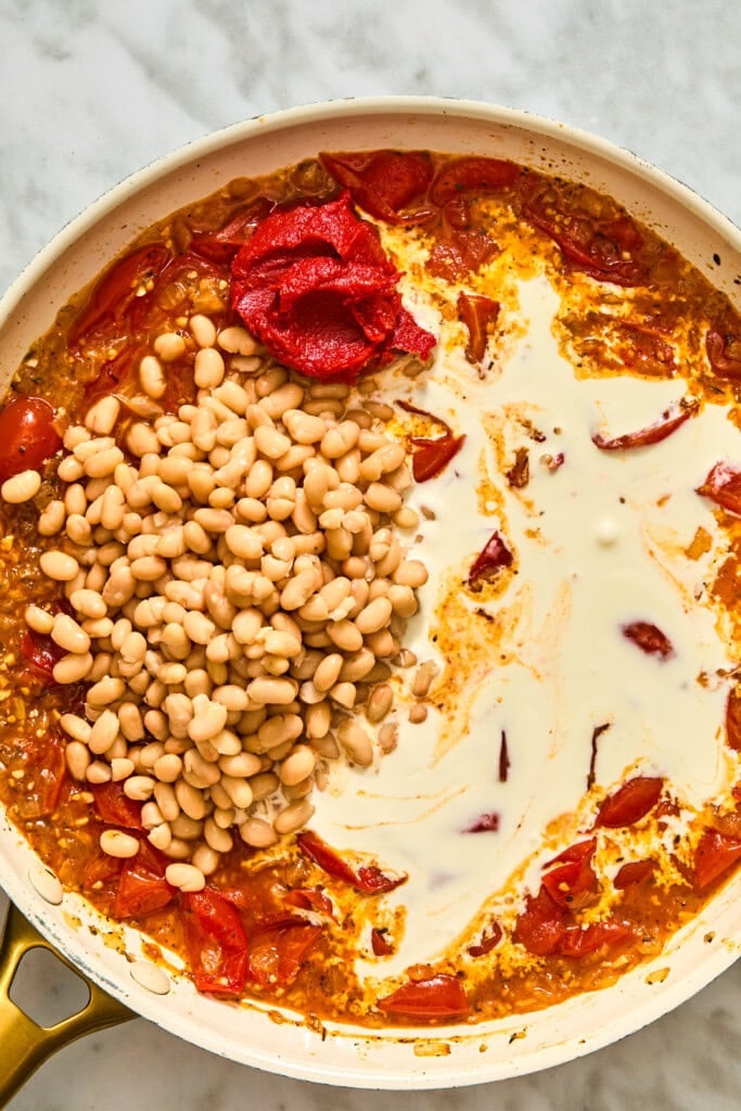 A skillet containing a sauce of cooked tomatoes, a pile of white beans, tomato paste, and a swirl of cream being prepared for creamy Tuscan white beans.