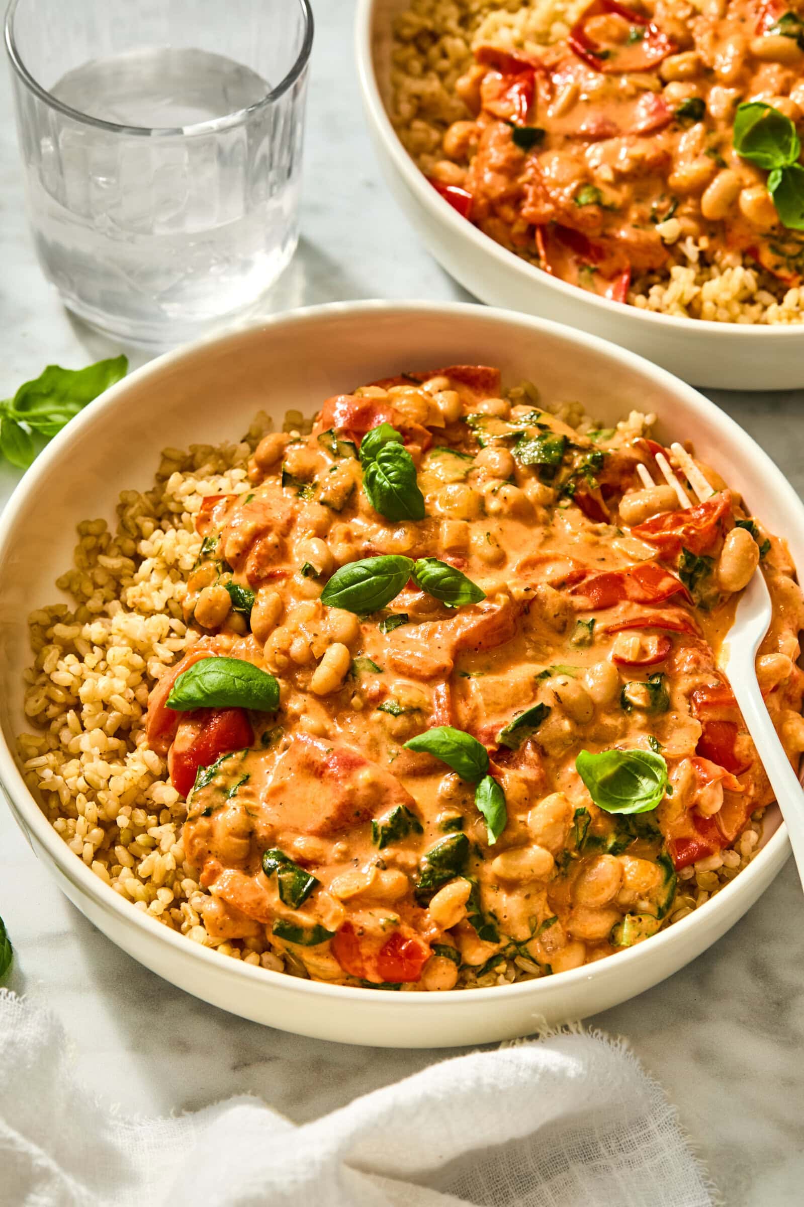 Two bowls of creamy Tuscan white bean and tomato stew served over brown rice, garnished with fresh basil leaves.