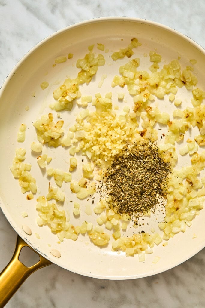 Sautéed diced onions, minced garlic, and a pile of dried herbs in a white skillet, the base for creamy tuscan white beans.