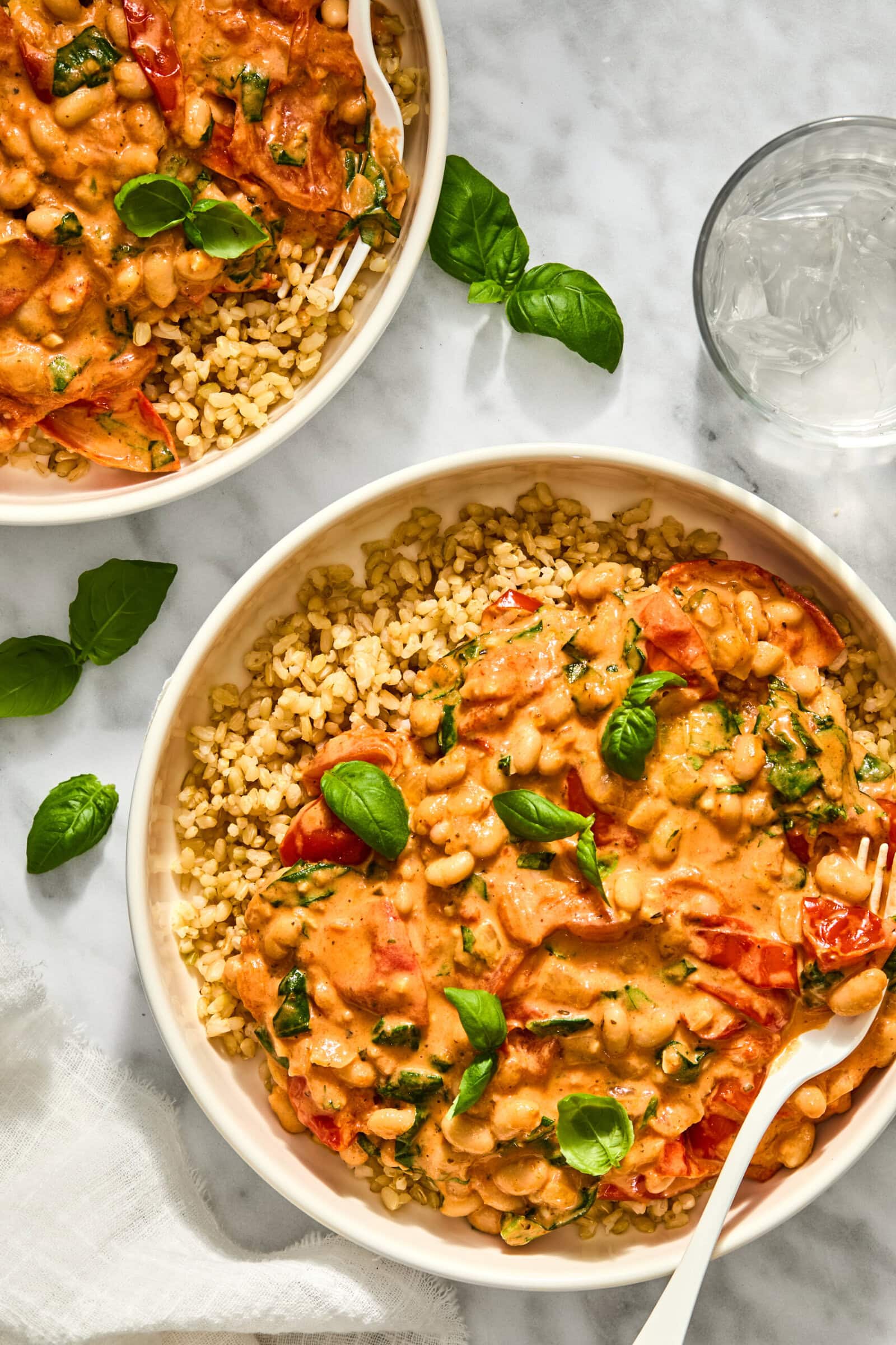Two bowls of creamy Tuscan white beans and greens served over brown rice, topped with fresh basil leaves.