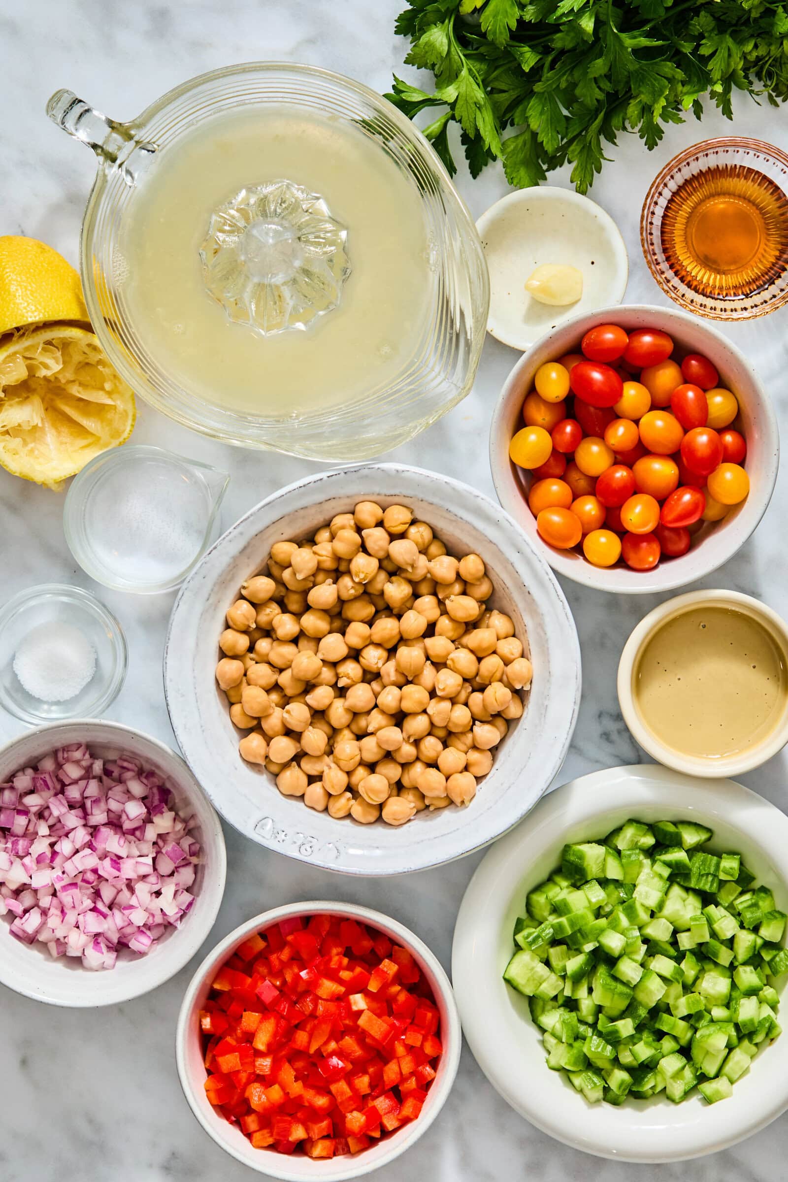 A top-down view of ingredients for a high-protein chickpea salad, including bowls of chickpeas, red onion, bell pepper, cucumber, tomatoes, and tahini, alongside a juicer with lemon juice, a garlic clove, oil, and parsley.