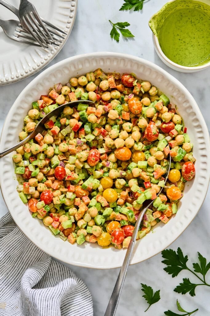 A large white bowl of high-protein chickpea salad with chopped tomatoes, cucumbers, and red onion, served with two spoons and a side of green dressing.