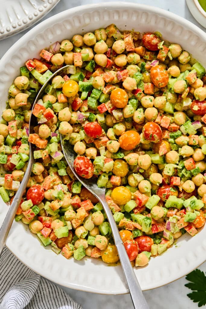 A large bowl of high-protein chickpea salad with diced cucumbers, tomatoes, and red onion, tossed with two serving spoons.