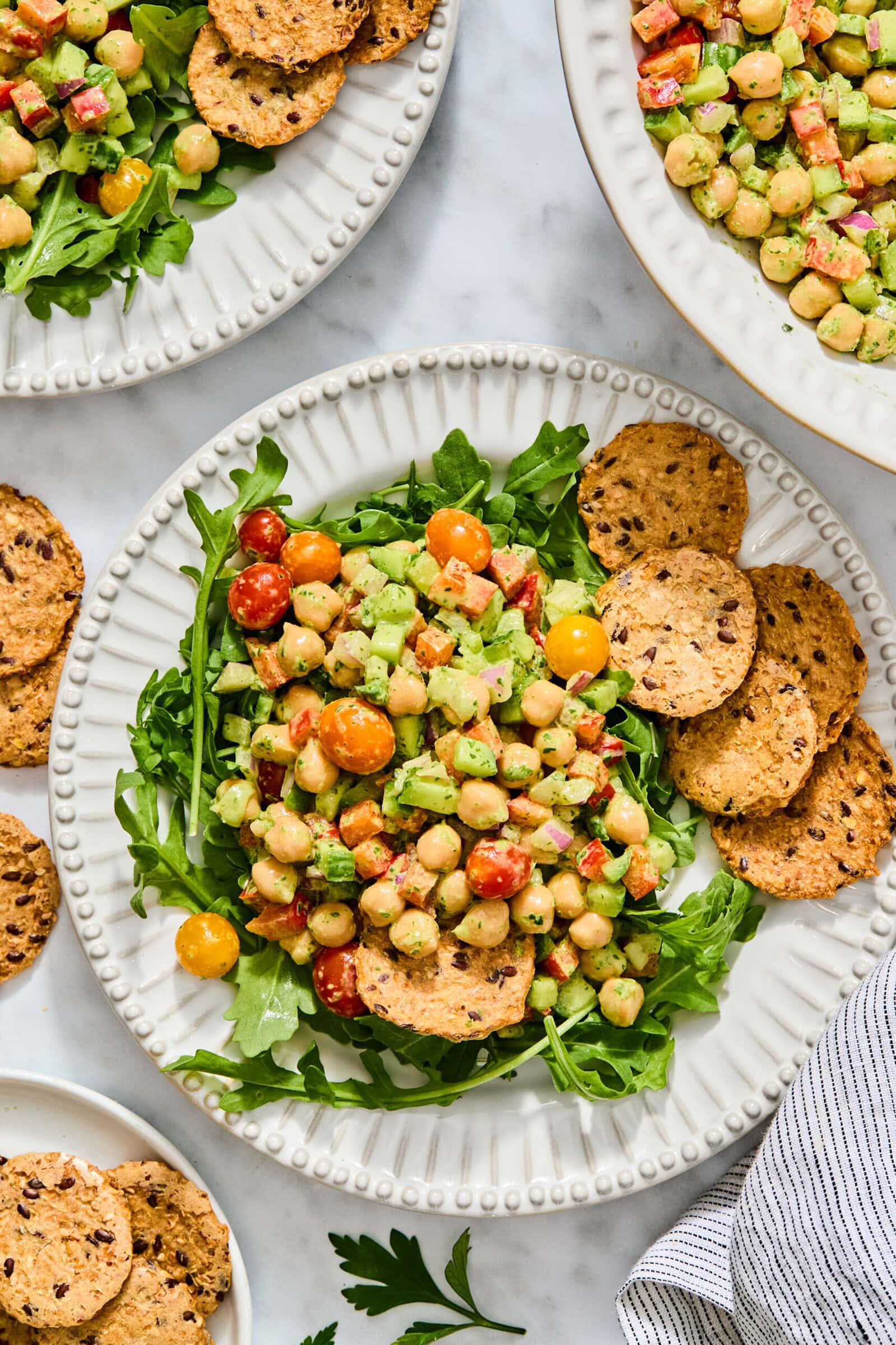 High-protein chickpea and vegetable salad served over arugula with a side of seeded crackers on a scalloped plate.