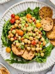 A bed of arugula topped with a high-protein chickpea and vegetable salad, served with several seeded crackers on a white scalloped plate.