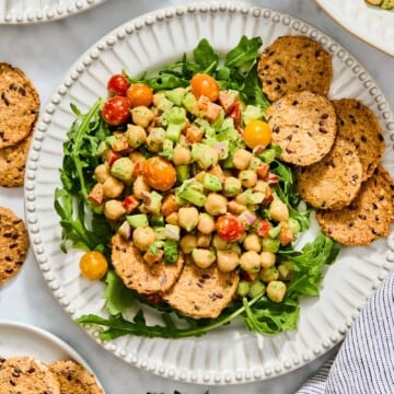 A bed of arugula topped with a high-protein chickpea and vegetable salad, served with several seeded crackers on a white scalloped plate.