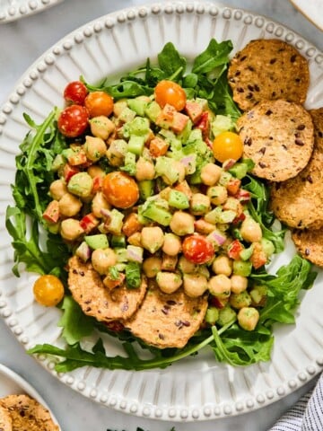 A bed of arugula topped with a high-protein chickpea and vegetable salad, served with several seeded crackers on a white scalloped plate.