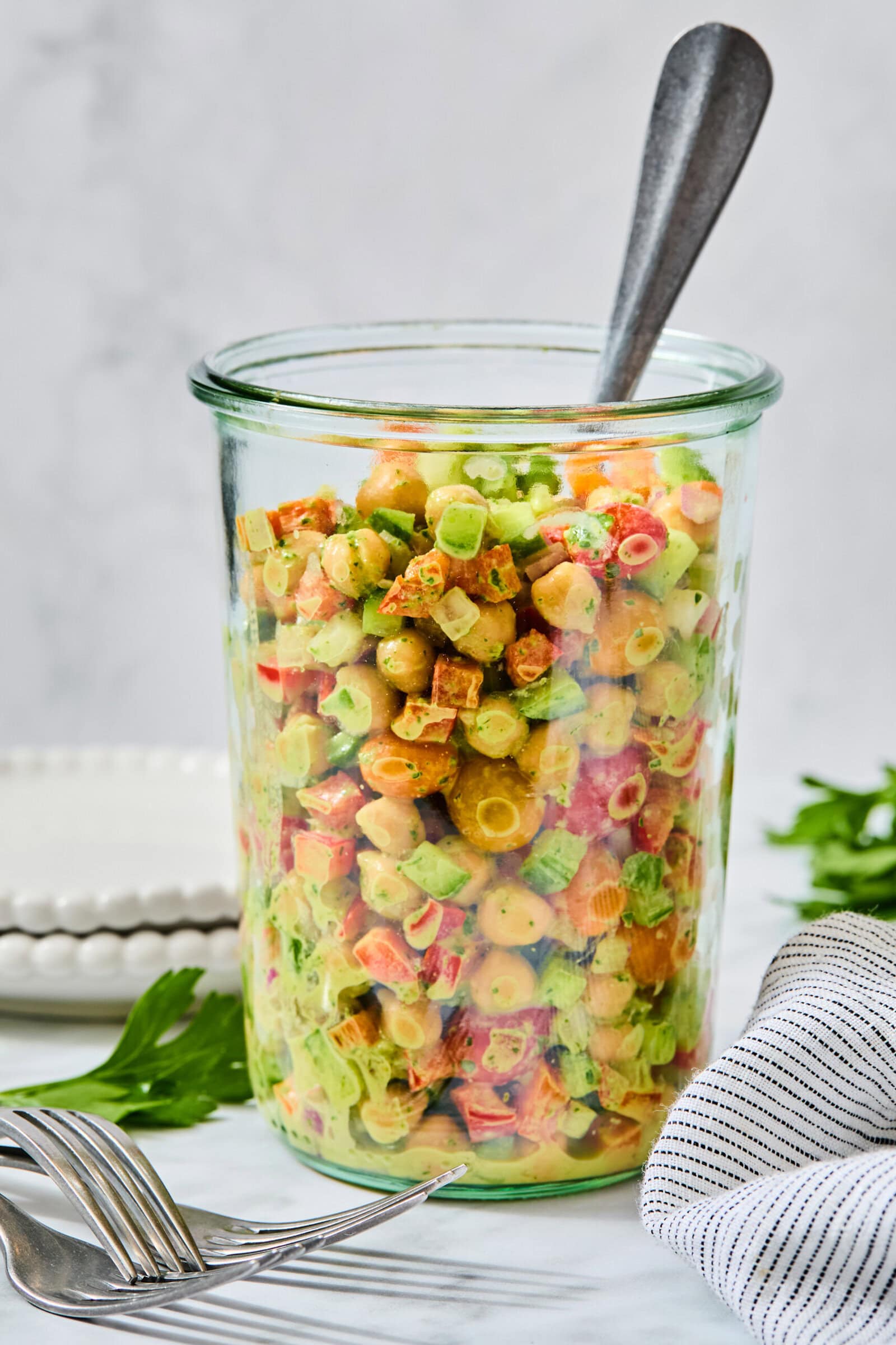 A glass jar filled with a high-protein chickpea salad, with a spoon resting inside and forks placed on the table nearby.