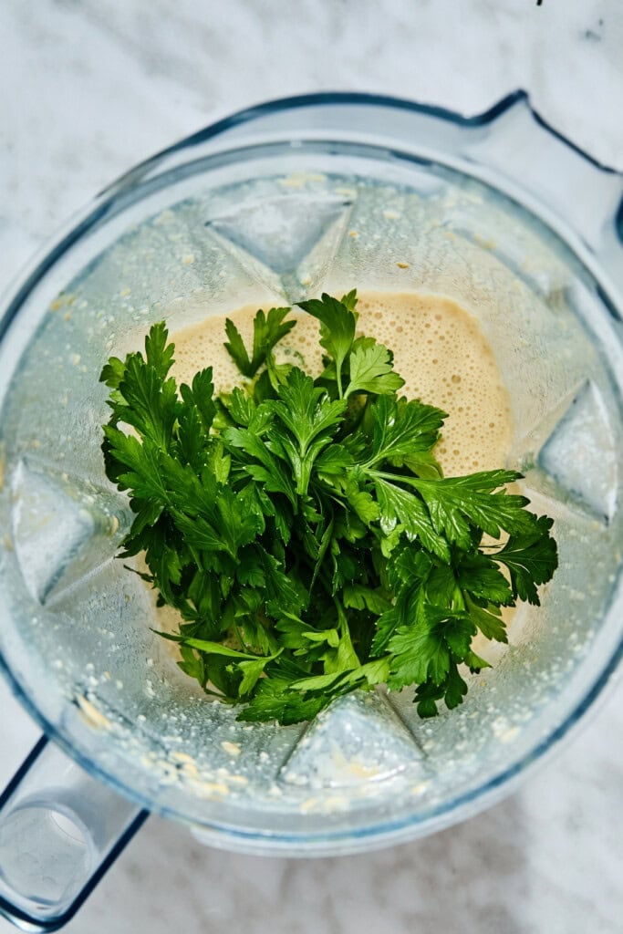 A bunch of fresh parsley sits in a blender jar filled with a creamy, pale yellow liquid base for a high-protein chickpea salad.