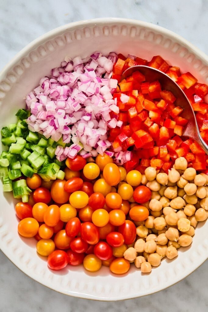 A white bowl filled with a high-protein chickpea salad containing sections of diced red onion, diced red bell pepper, chopped cucumber, chickpeas, and a mix of yellow and red cherry tomatoes, with a serving spoon resting in the bowl.