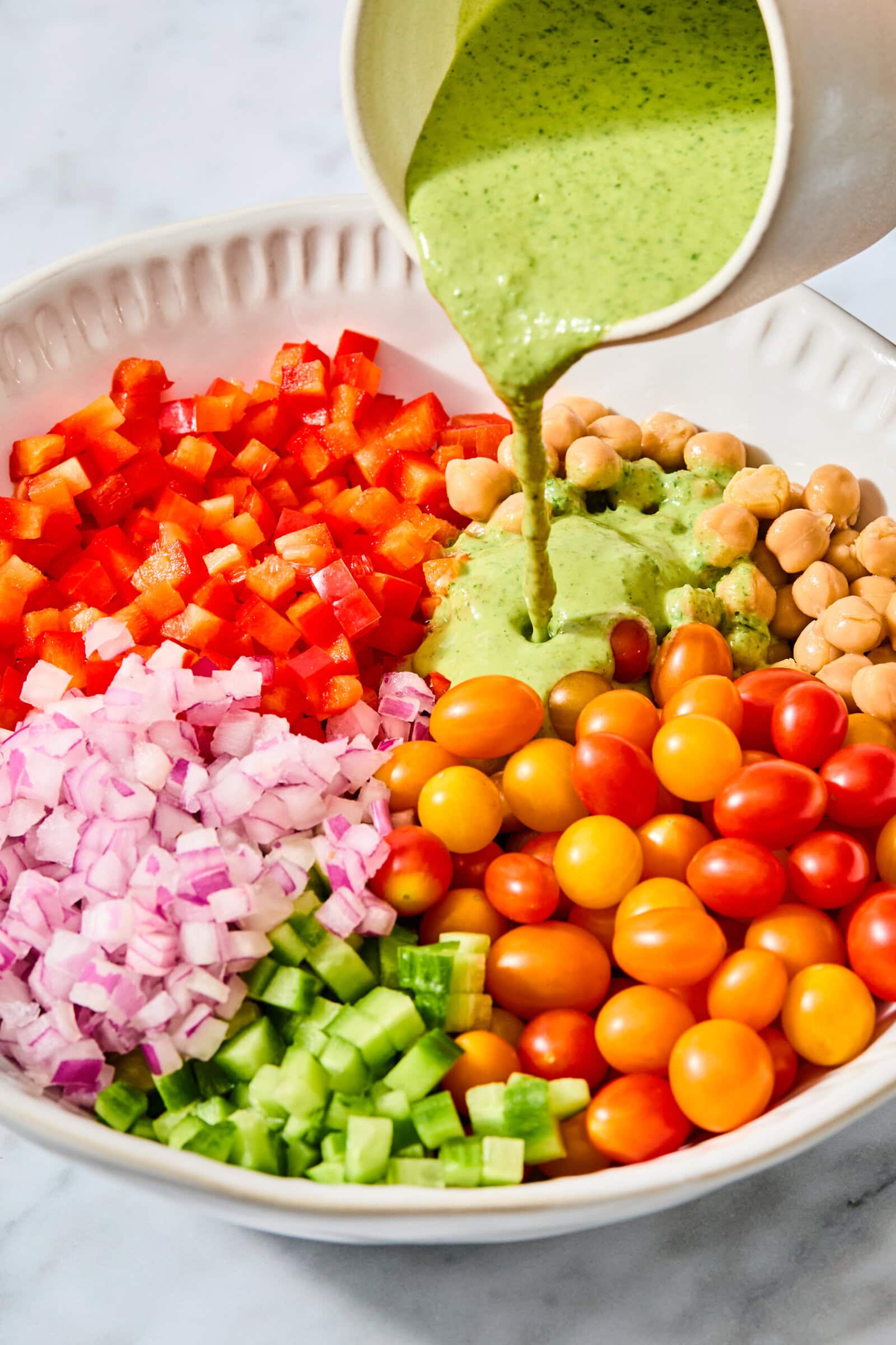 Green dressing is poured from a pitcher into a high-protein chickpea salad containing chopped red peppers, red onions, cucumbers, and cherry tomatoes.