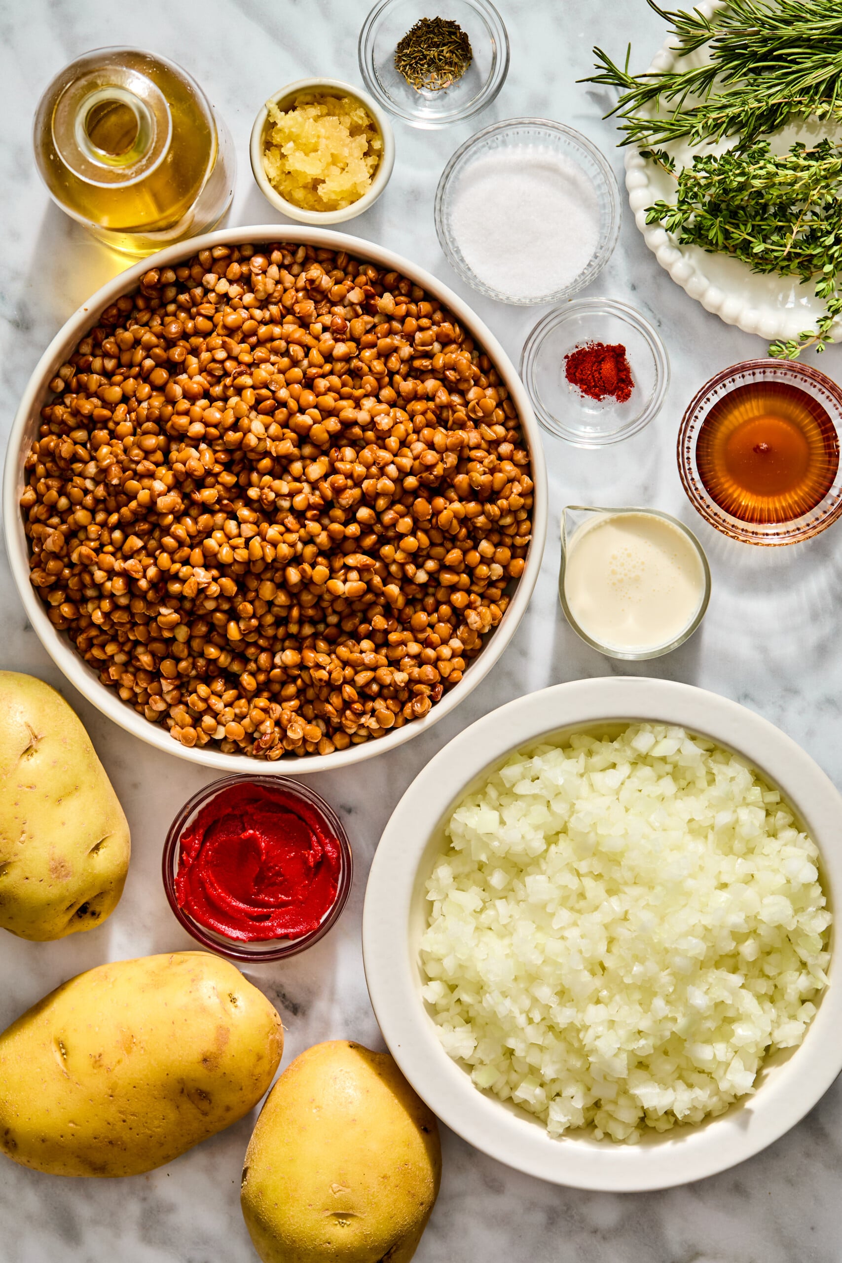 A large bowl of cooked lentils next to a bowl of chopped onions, three raw potatoes, and various small bowls containing olive oil, minced garlic, spices, tomato paste, cream, and fresh herbs.